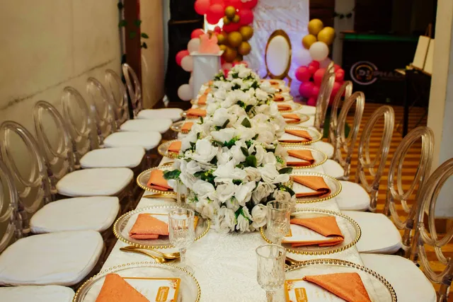 Mesa de celebración decorada con flores blancas, vajilla dorada y globos al fondo